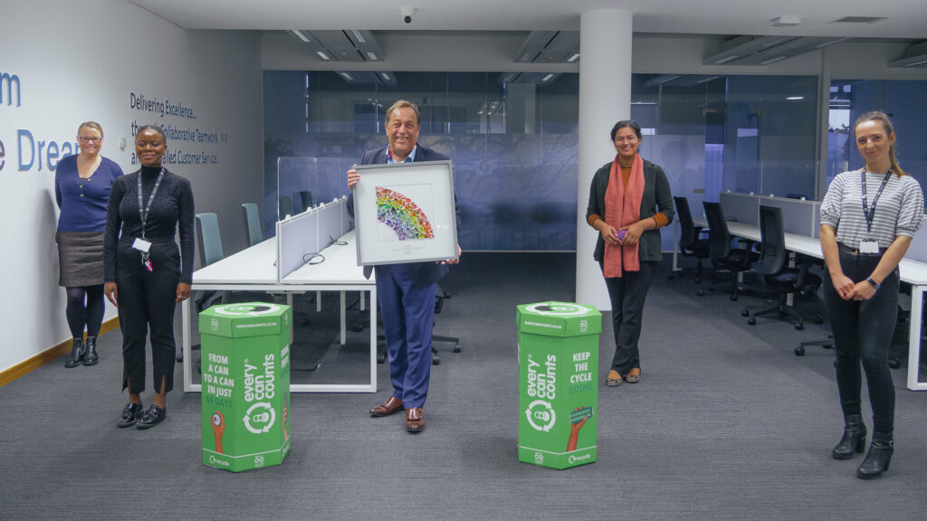 Socially distanced photoshoot of Subfero team in office setting with two Every Can Counts collection boxes between each person. In the middle, a Subfero team member holds artwork made from recycled cans which was presented as a gift to celebrate the recycling efforts of the company who have succeeded in promoting recycling in their workplace. 