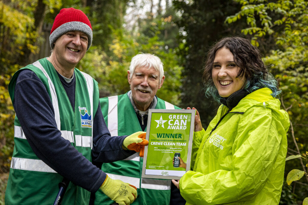 On the left of the photo is David (chair of Crewe Clean Team) who is being presented with a Green Can Award Certificate by Phoebe (Programme Assistant at Every Can Counts). Behind them both is Arthur, another member of Crewe Clean Team. Everyone in the photo is laughing and happy. 
