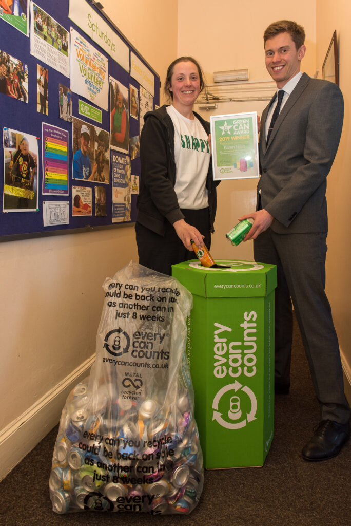 Karen Nash from Foil For Snappy poses with Chris Latham-Warde from Every Can Counts behind a drink can collection box. They both hold a Green Can Award certificate and are each recycling an empty drink can. 