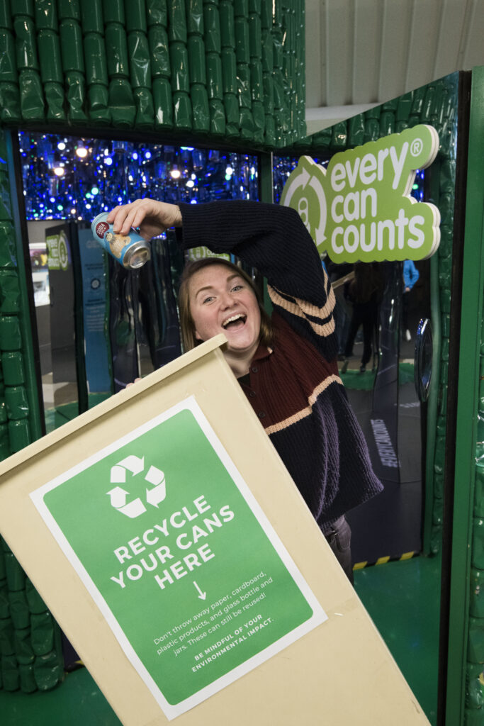Less Waste Laura stands in front of the #EveryCanCounts Infinity Room, holding an empty can over a recycling bin. She is smiling and is enjoying her visit. 